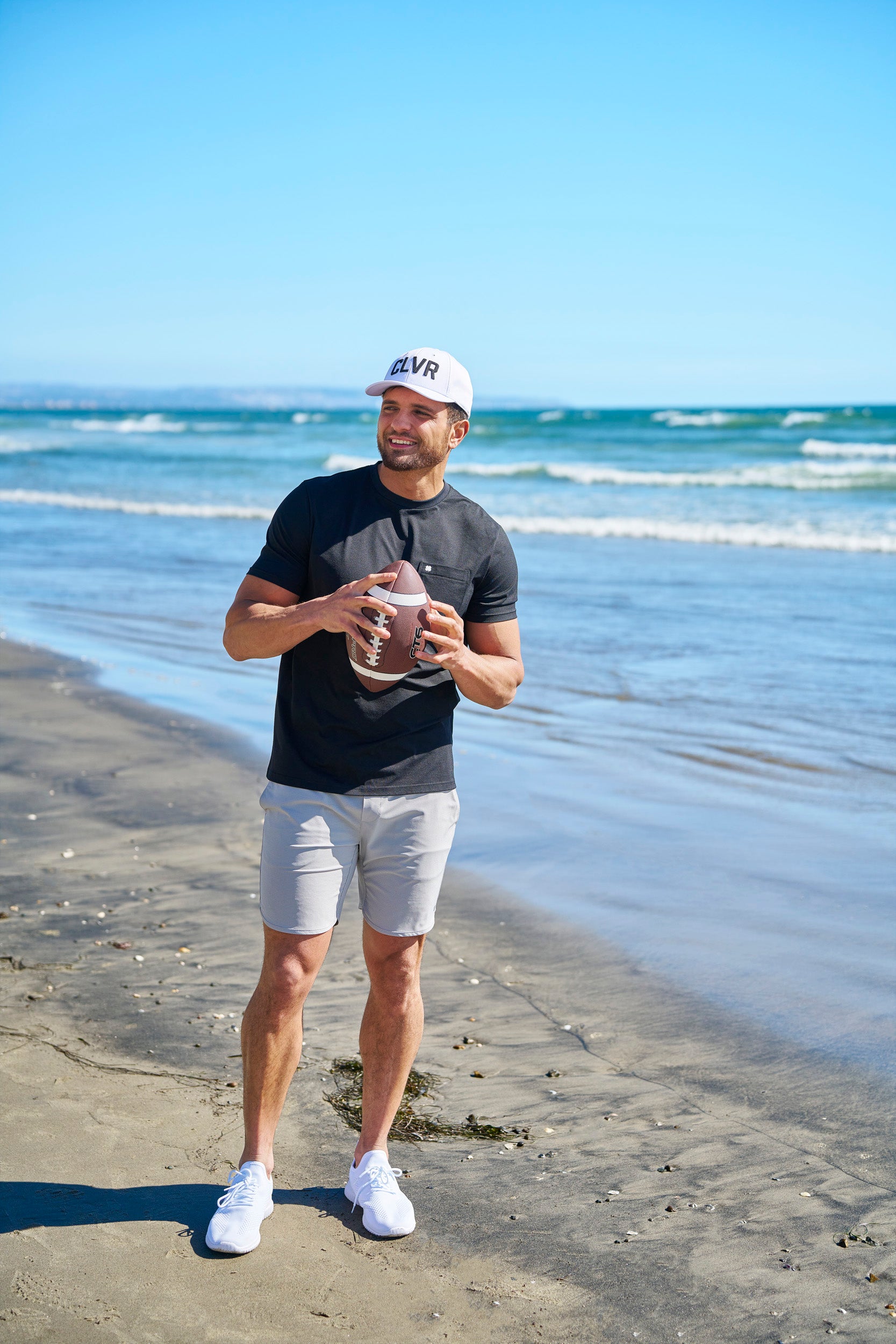 Man holding american football at the beach wearing CLVR Black Clover Live Lucky hat.