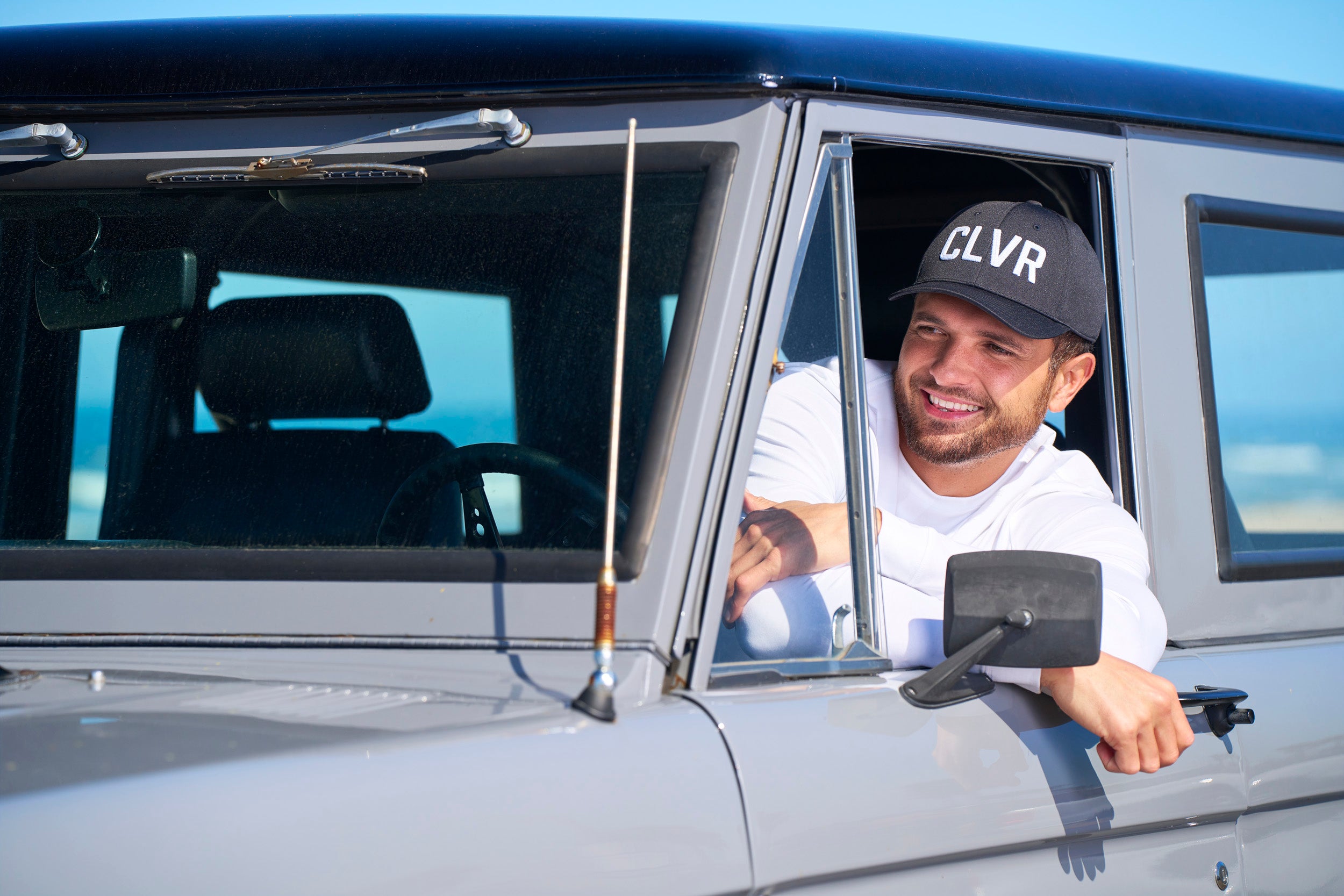Man leaning out the window of his car wearing a Black Clover Live Lucky CLVR hat in white.
