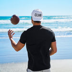 Man tossing an american football at the beach wearing a Black Clover Live Lucky CLVR hat in white.