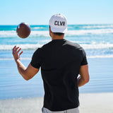 Man tossing an american football at the beach wearing a Black Clover Live Lucky CLVR hat in white.